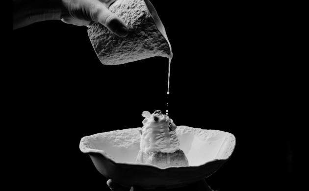 Dressage d’un dessert raffiné avec une sauce versée délicatement depuis un petit pichet, en noir et blanc, sur fond sombre.