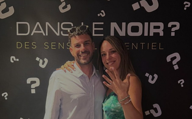 Un couple souriant après une demande en mariage réussie chez Dans le Noir ? Marseille, devant le mur aux points d'interrogation iconiques.