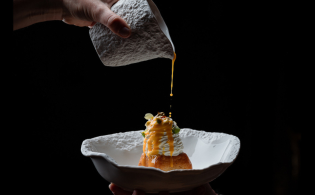 Dressage gastronomique d'un plat raffiné avec une main versant délicatement une sauce dorée sur une préparation dans une assiette texturée blanche, sur fond noir.