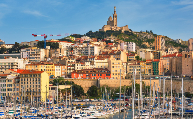 Vue panoramique du Vieux-Port de Marseille avec ses nombreux voiliers, en arrière-plan la basilique Notre-Dame-de-la-Garde dominant la ville sous un ciel bleu.